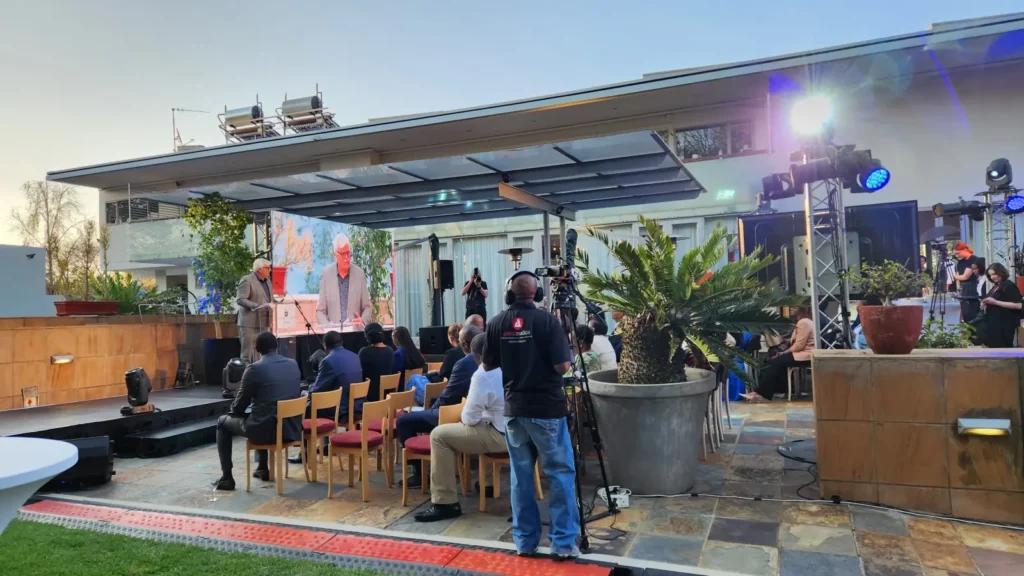 Camera operator filming an outdoor product launch on a rooftop patio, with two speakers on stage and a small seated audience