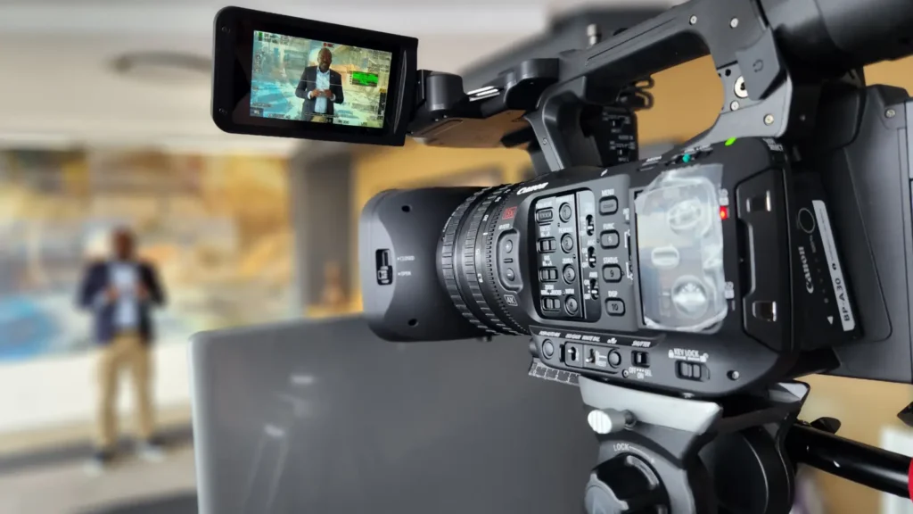 Close-up of a Canon broadcast camera with a presenter visible on the flip-out screen, filming indoors for a webinar