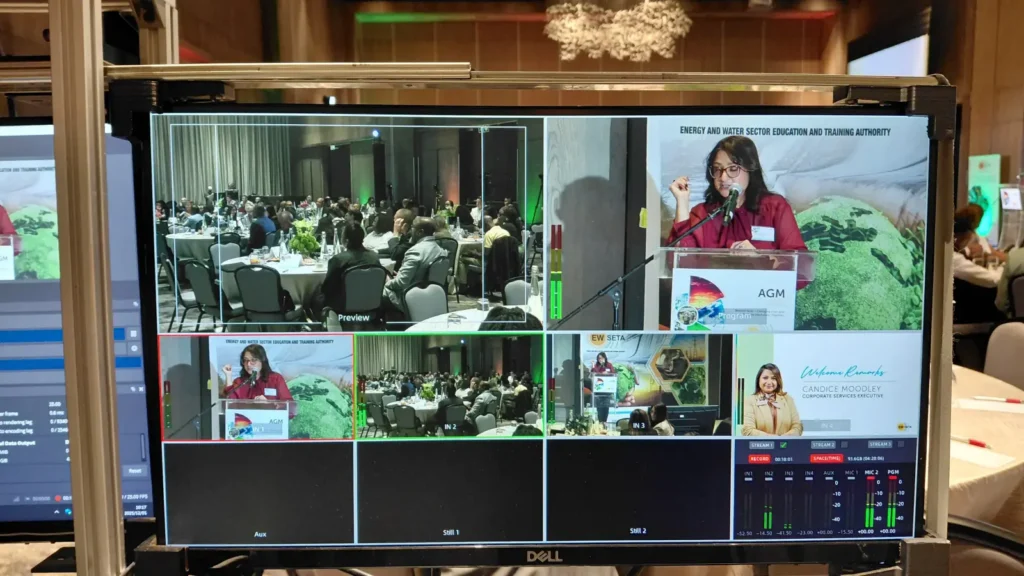 Broadcast production monitor showing a live AGM stream, with a speaker at the podium, audience shots, and a welcome remarks graphic for the corporate services executive.