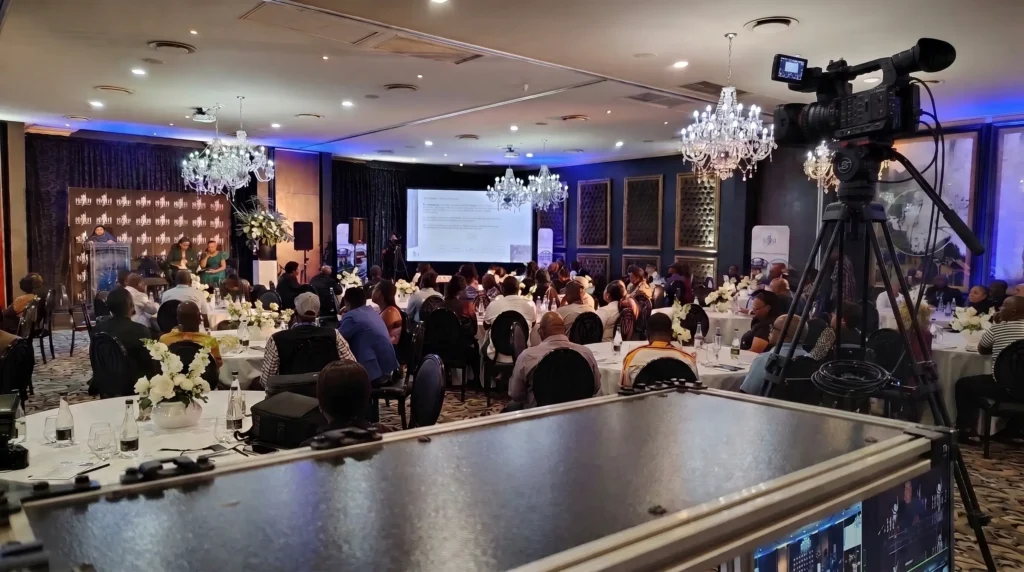 Camera and monitors overlooking a chandelier-lit conference room, with a panel discussion on stage and guests seated at round tables