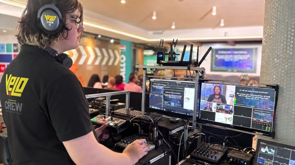 Production crew member in a branded shirt operating a switcher and mixing desk, with monitors showing a live speaker feed and captions