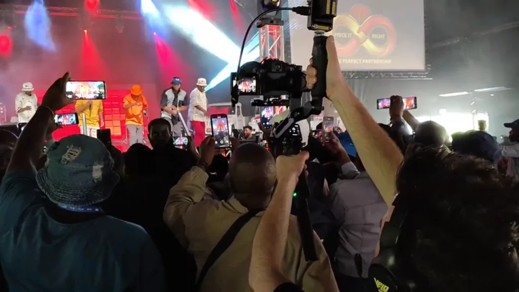 Camera operator holding a gimbal camera above a crowd at a live concert, filming performers on a stage lit with red and blue lights.