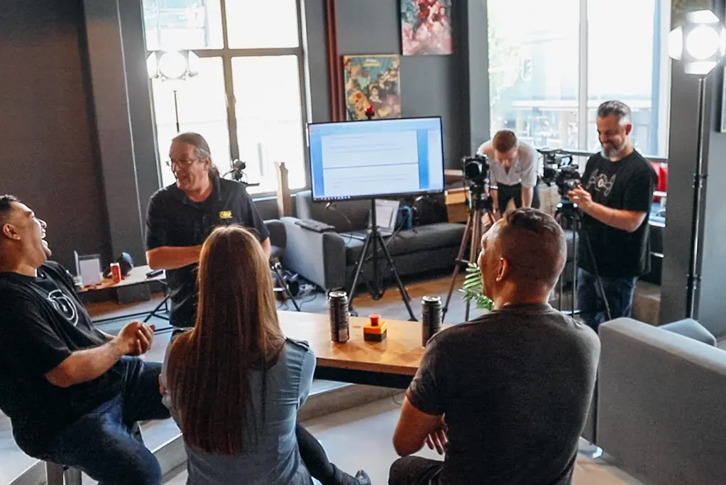Group filming a lounge-style discussion indoors, with people seated around a coffee table and two crew members adjusting cameras behind them