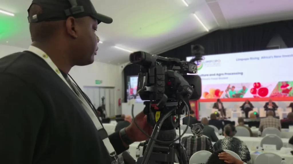 Camera operator filming a panel discussion at a conference, with a large screen behind the stage reading "Limpopo Rising"
