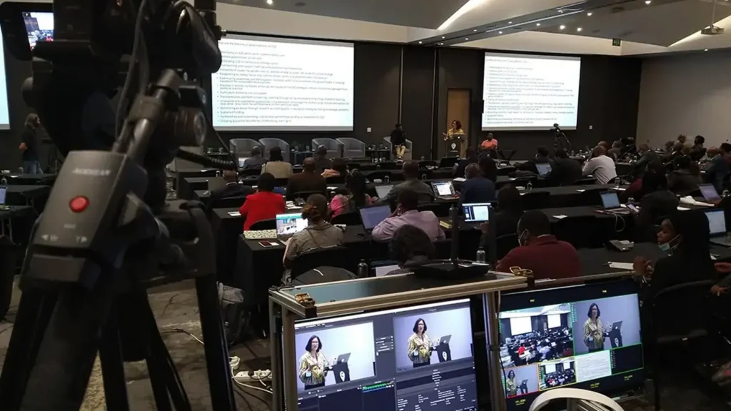 Camera and production monitors at the back of a large conference room, with a speaker at the podium and delegates seated at rows of tables.