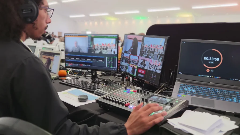 Technician operating a video switcher and multi-camera monitors during live AGM production, with an on-air indicator visible on screen.