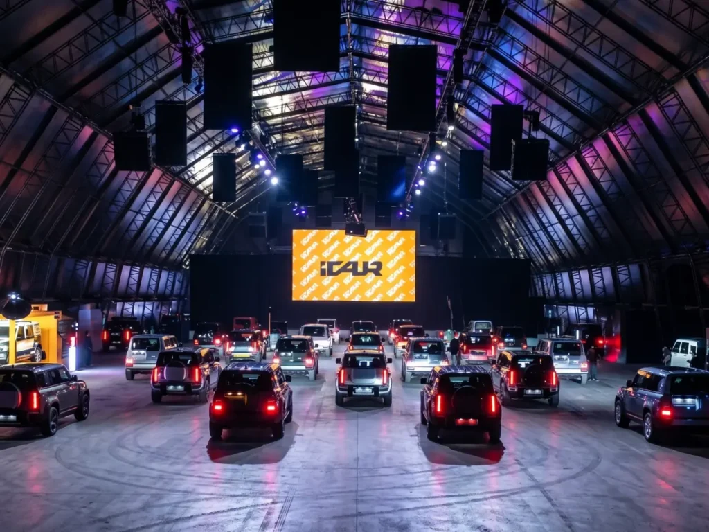 Rows of SUVs parked inside a large industrial hangar venue, facing a big screen with orange ICAUR branding, under purple and blue stage lighting with rigged speakers overhead.
